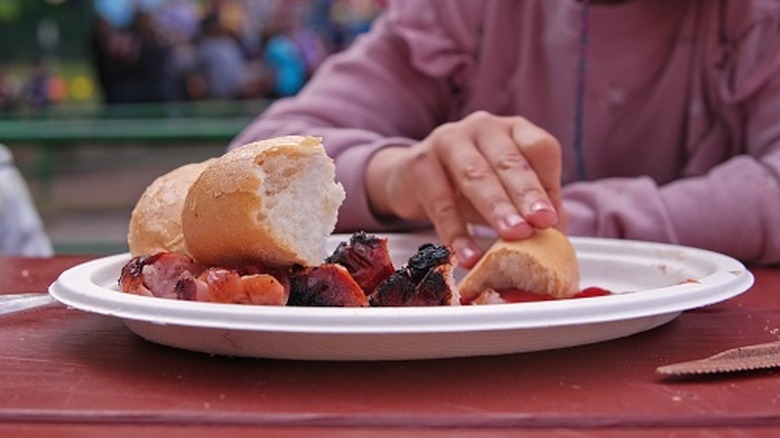 A girl picks up food from a disposable plate on a red picnic table outside.