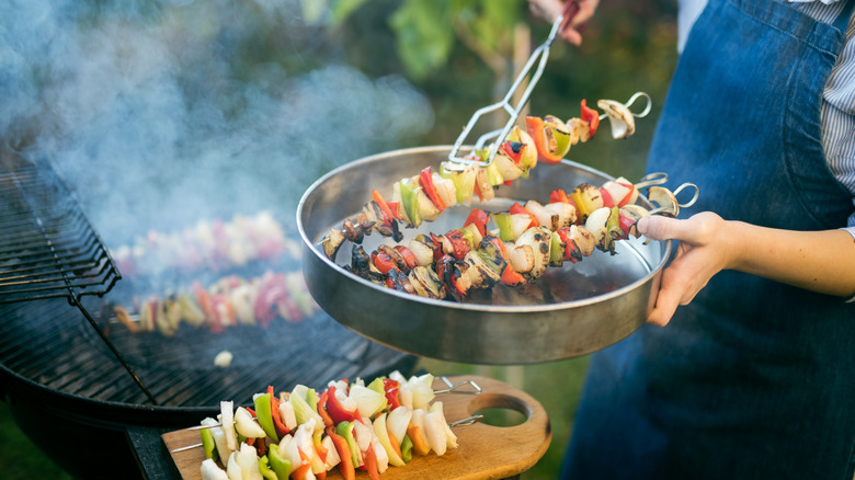 A person in a blue apron removing vegetable skewers from a  smoking grill