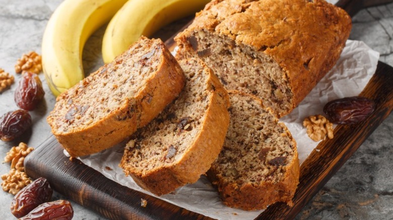 A sliced loaf of banana bread next to bananas, dates, and walnuts