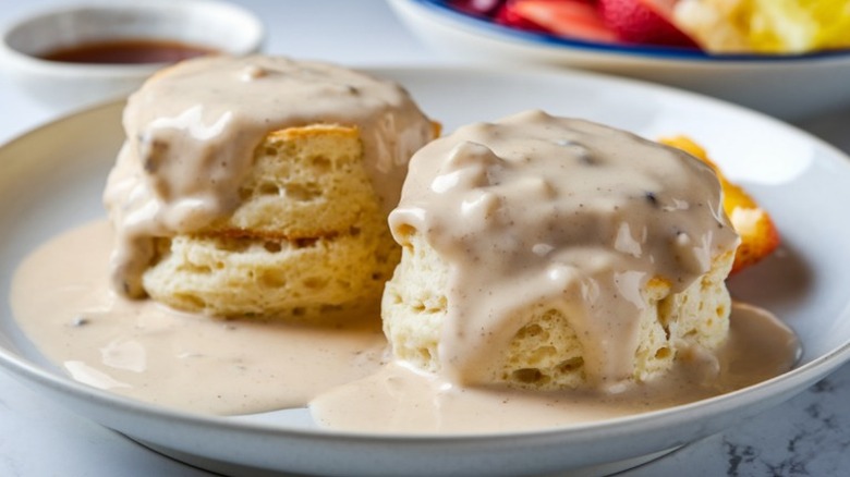 Two biscuits with gravy on a plate with fruit in background