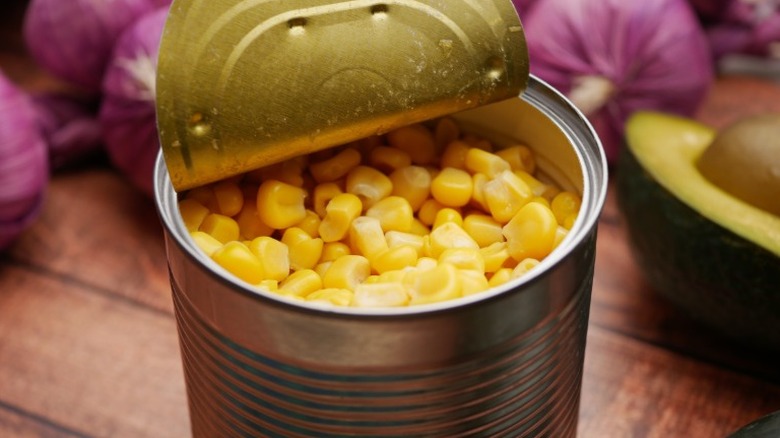 An opened can of corn on a wooden table with onions and avocados in background
