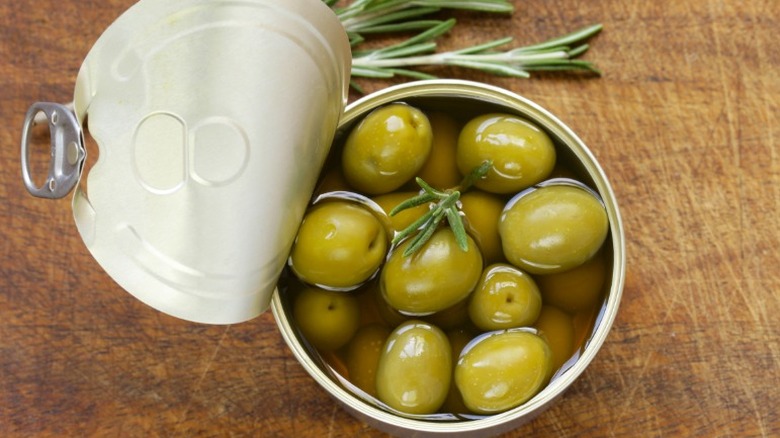An overhead shot of an open can of green olives on a wooden counter