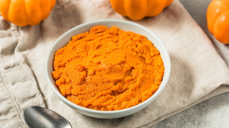 A bowl of pumpkin puree sits on a beige cloth with whole pumpkins and silverware.