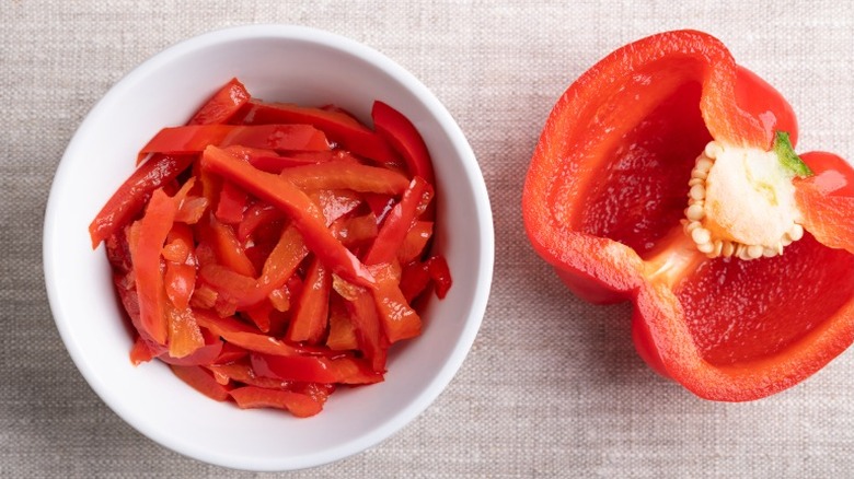 A bowl of sliced red peppers next to a halved red bell pepper
