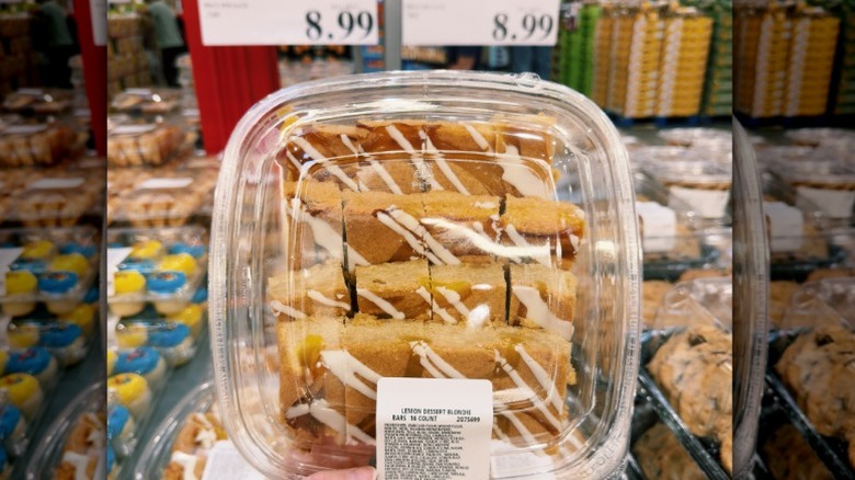 A hand holding a plastic tub of Costco lemon blondie dessert bars in the store.