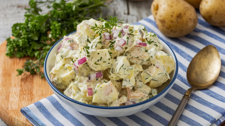 A bowl of potato salad styled on a table with potatoes, parsley, and a dish cloth