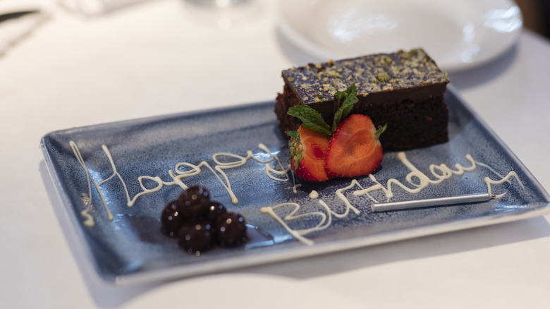 A blue rectangular plate rests on a table with a brownie and fruit surrounding it. Happy Birthday is written in icing on the plate.