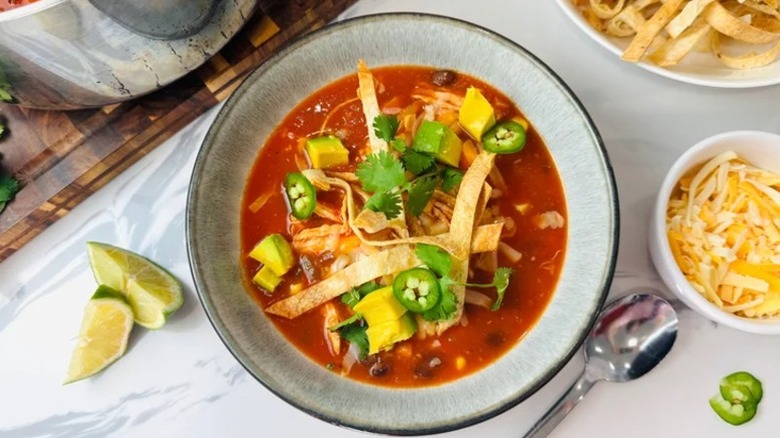 A bowl of chicken tortilla soup topped with cheese, jalapeños, avocado, and tortilla strips on top