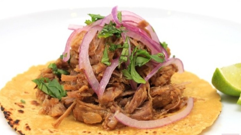 Open-faced carnitas tacos in corn tortillas topped with red onions and cilantro, lime wedges on the side