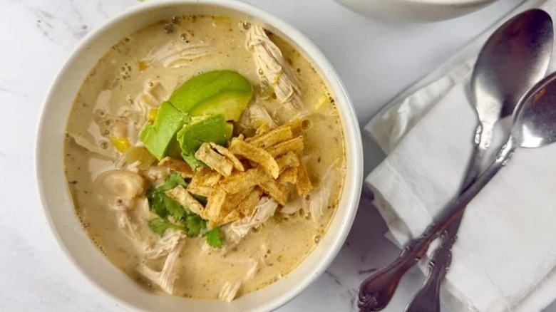 Bowl of white chicken chili topped with avocado and tortilla strips with spoons and napkins to the side