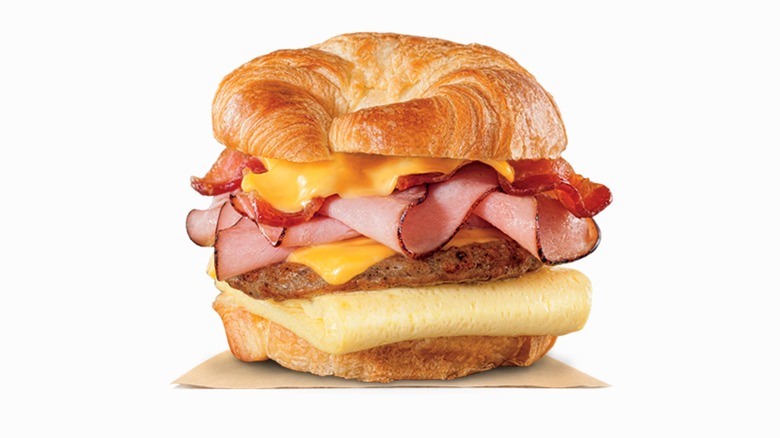 A Burger King Croissant-style breakfast sandwich with egg, sausage, cheese, ham, and bacon, sitting on a piece of waxed paper on a white background