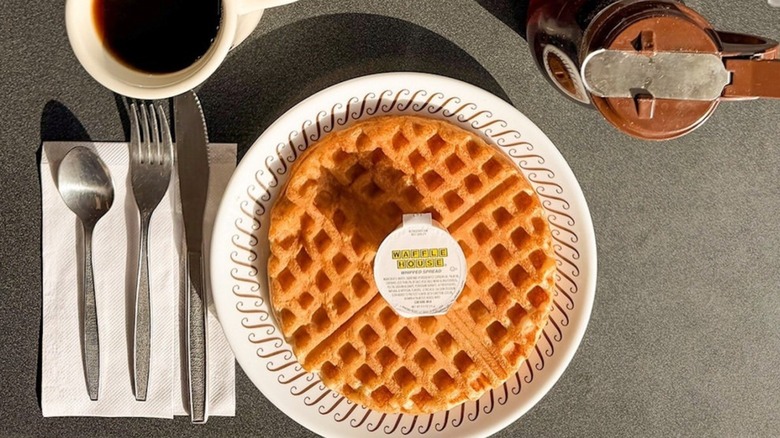 Top view of Waffle House waffle with packet of butter on top alongside silverware, cup of coffee, and syrup pitcher