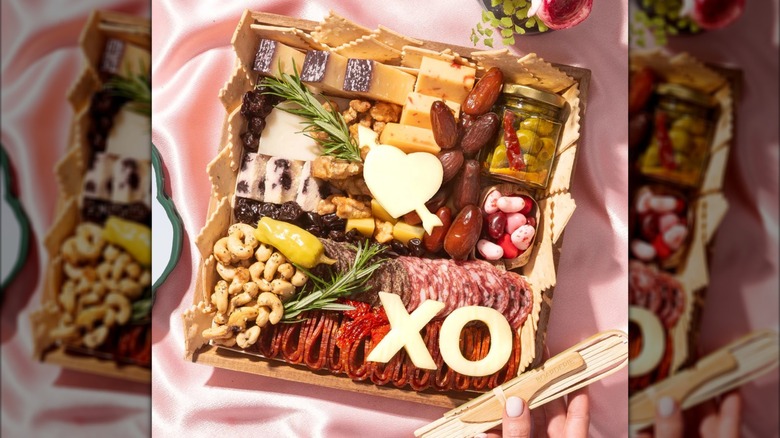 Valentine's Classic Cheese and Charcuterie Board with jars of olives, cut out cheese, cashews, dates, and meat from Boarderie on top of a pink satin cloth