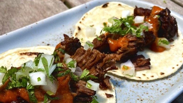 Two open soft tacos topped with slow cooked beef, tomato sauce, diced onions, and cilantro