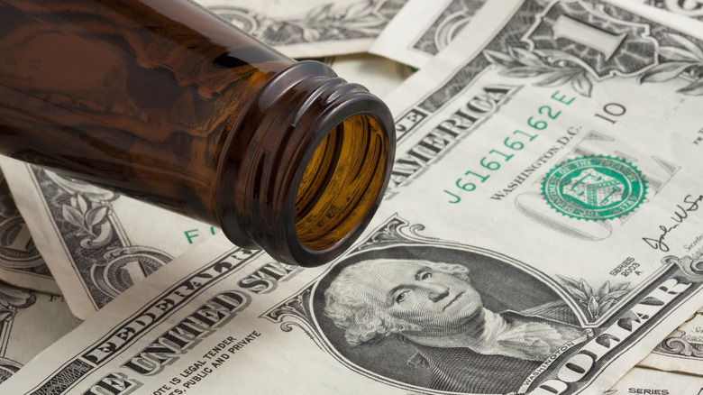 An empty beer bottle on top of a stack of dollar bills.