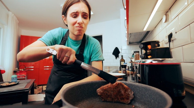 Person with disappointed facial expression holding steak in pan with tongs