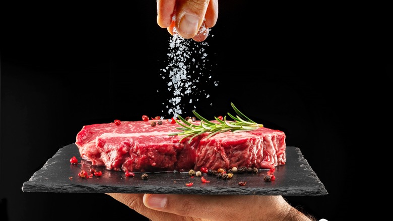 Person sprinkling salt onto red meat on black surface