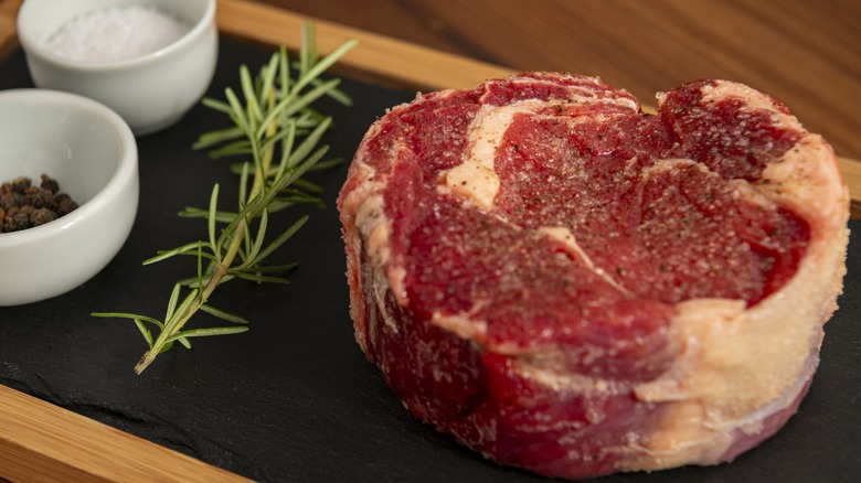 Raw seasoned steak sitting on black surface next to rosemary sprig and white bowls with salt and pepper