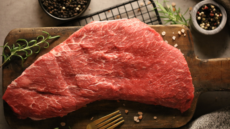 A whole, raw flat iron steak on a wooden cutting board with seasonings scattered around.