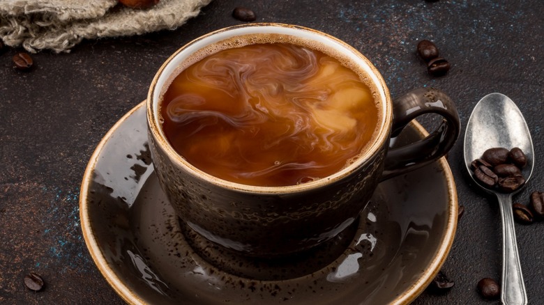 Coffee with creamer in dark cup on saucer with spoon and coffee beans nearby
