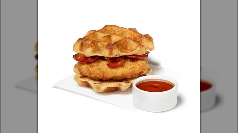The Chicken & Waffles Breakfast Sandwich from Chick-fil-A with bacon on it and a cup of syrup beside it.