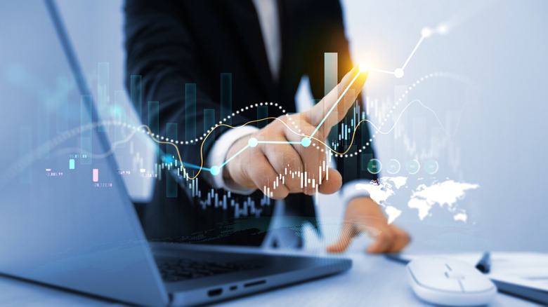 A man's hand points to an upward trending stock market graph with a computer in the background