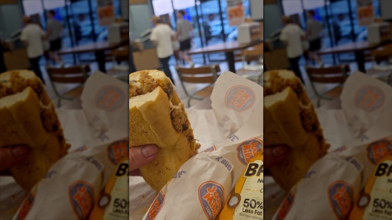 A hand holds a Jersey Mike's sub sandwich at a table inside the restaurant
