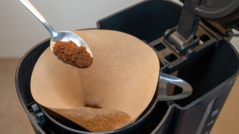 Spoon pouring ground coffee into a brown paper filter in a drip coffee machine