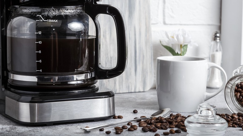 Drip coffee maker with nearly full pot of coffee next to white ceramic mug and scattered whole coffee beans