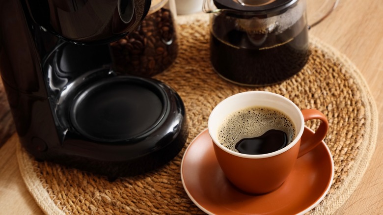 Black coffee in a ceramic cup with a saucer next to a coffee maker and whole beans