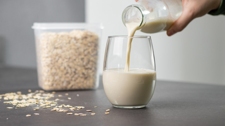 A hand pouring oat milk out of a glass jar into a drinking glass with container of dried oats in the back