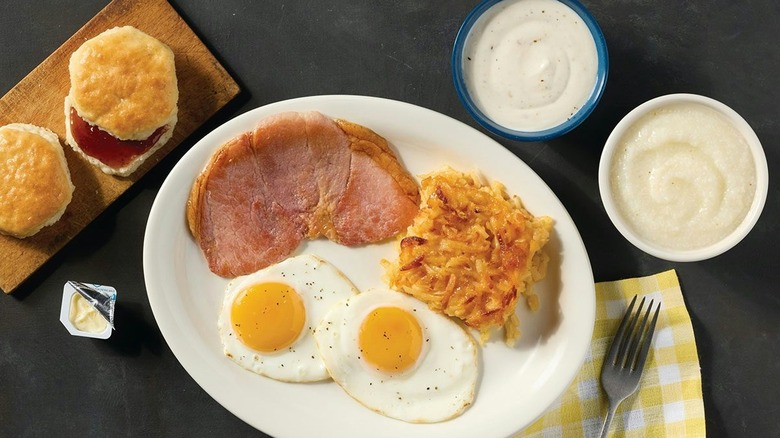 A plate of eggs, hashbrowns, and pork on a black tabletop with dips and two biscuits with jelly around it.