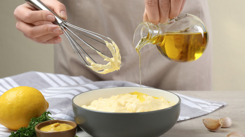 Hands holding whisk and bottle of olive oil that's being drizzled into a large bowl of homemade mayonnaise