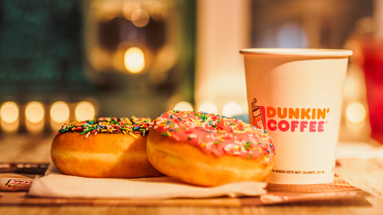 Two iced donuts with sprinkles, one pink and one chocolate, beside a take out Dunkin' coffee cup in front of a soft focus background