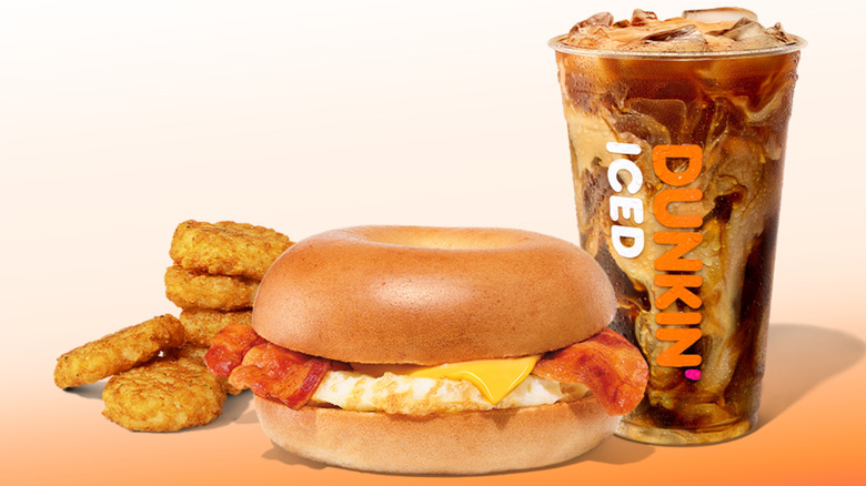 A large iced coffee in a Dunkin' cup alongside an egg, cheese, and bacon bagel and a pile of small hash browns
