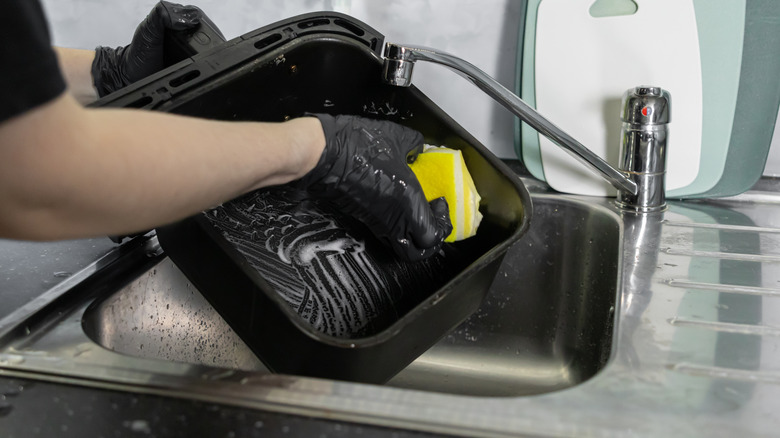 person wearing black rubber gloves using a yellow sponge to scrub a black air fryer basket held over a stainless steel kitchen sink