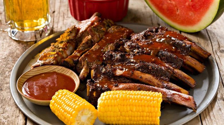 Platter full of smoked ribs, corn, spicy sauce, a bowl of baked beans, watermelon, and a mug of beer on a wood table.