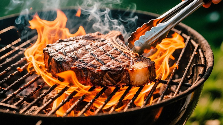 Tongs reaching for T-bone steak on charcoal grill with flames rising