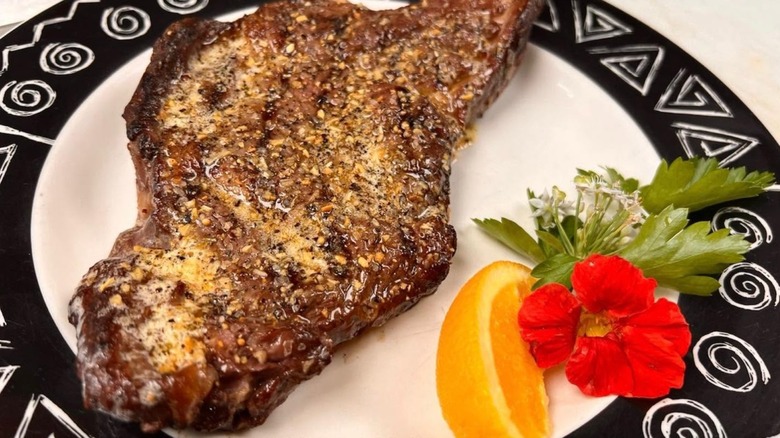 A bone-in bison ribeye on a white plate with a black and white patterned border with an orange wedge and a red flower garnish.