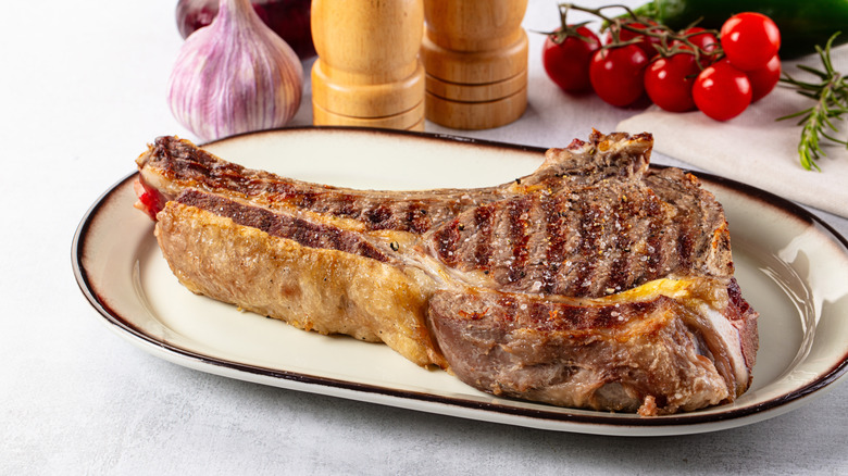A bone-in ribeye steak on an oval white plate with a brown rim posed in front of wooden salt and pepper grinders, a shallot, and cherry tomatoes on the vine