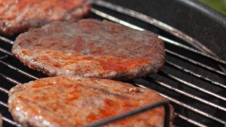 Several burger patties glazed with sauce cooking on an outdoor charcoal grill