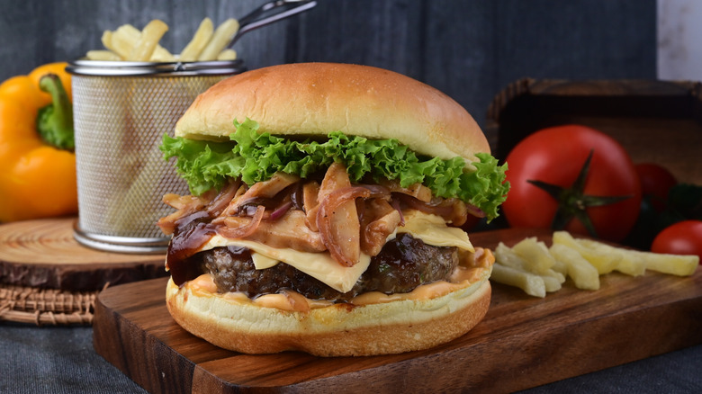 A burger with Swiss cheese, mushrooms, and lettuce on a wooden board with fries and whole tomatoes behind it