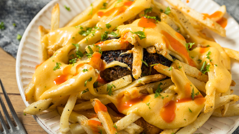 A horseshoe sandwich with a burger patty on a white plate buried under fries and topped with cheese sauce and hot sauce