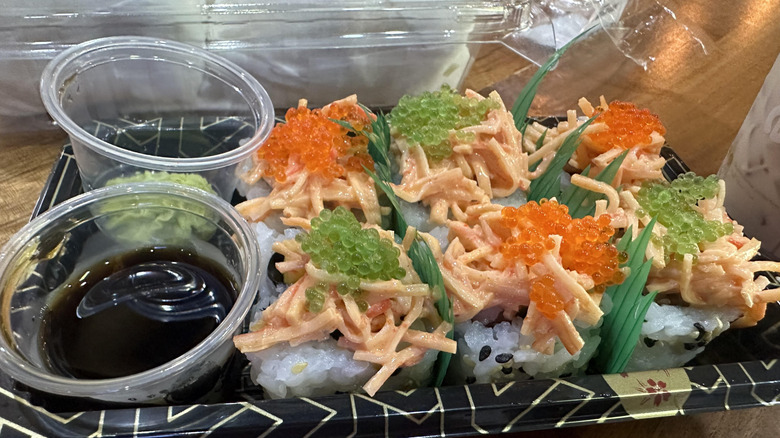 A take-out container of sushi with fish eggs on top and sauces on the side.