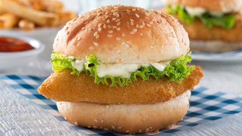 A fried fish sandwich with lettuce and sauce in front of a plate of fries and another sandwich.