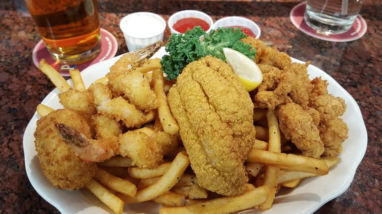 A white plate piled with fries, fried fish, and fried shrimp on a red table top with a beer visible beside the dish