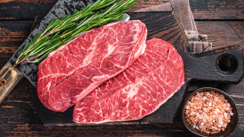 A pair of raw flat iron steaks stacked next to each other near some rosemary and a bowl of salt.
