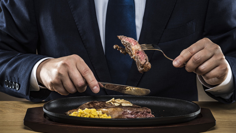 A man in a business suit sitting down and eating a steak.
