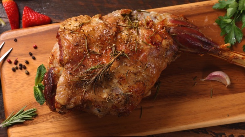 A roast leg of lamb on a wooden cutting board with herbs.