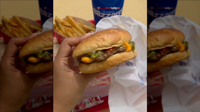 A hand holds a cheeseburger from Dairy Queen in front of fries and a Blizzard
